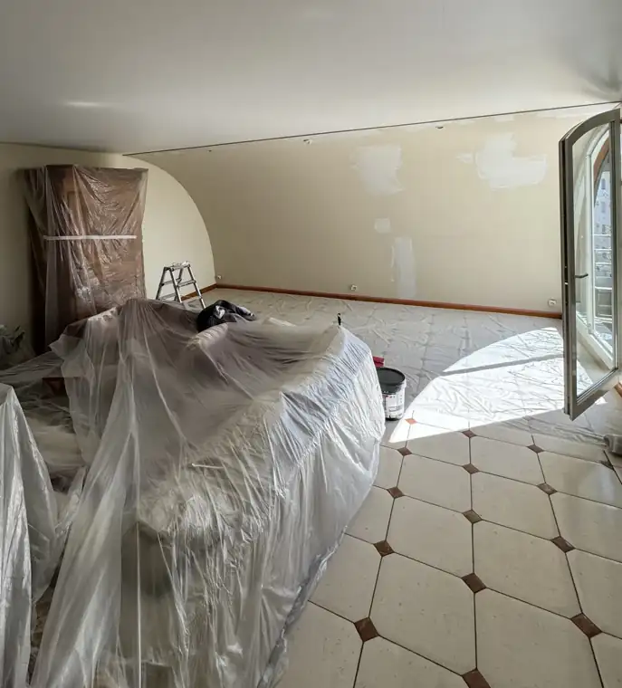 Le même séjour pendant le chantier : meubles regroupés au centre sous bâche, armoire emballée, sol protégé, rebouchages en cours et porte-fenêtre ouverte pour aérer